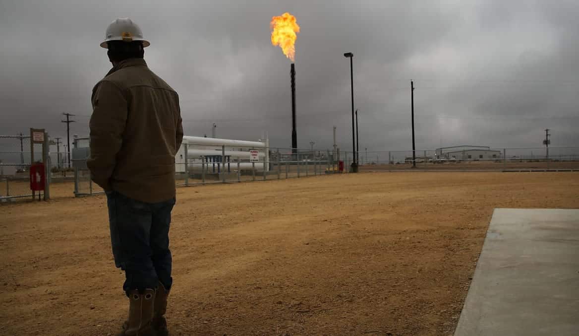 A man stands in a natural gas plant as gas is flared in the distance